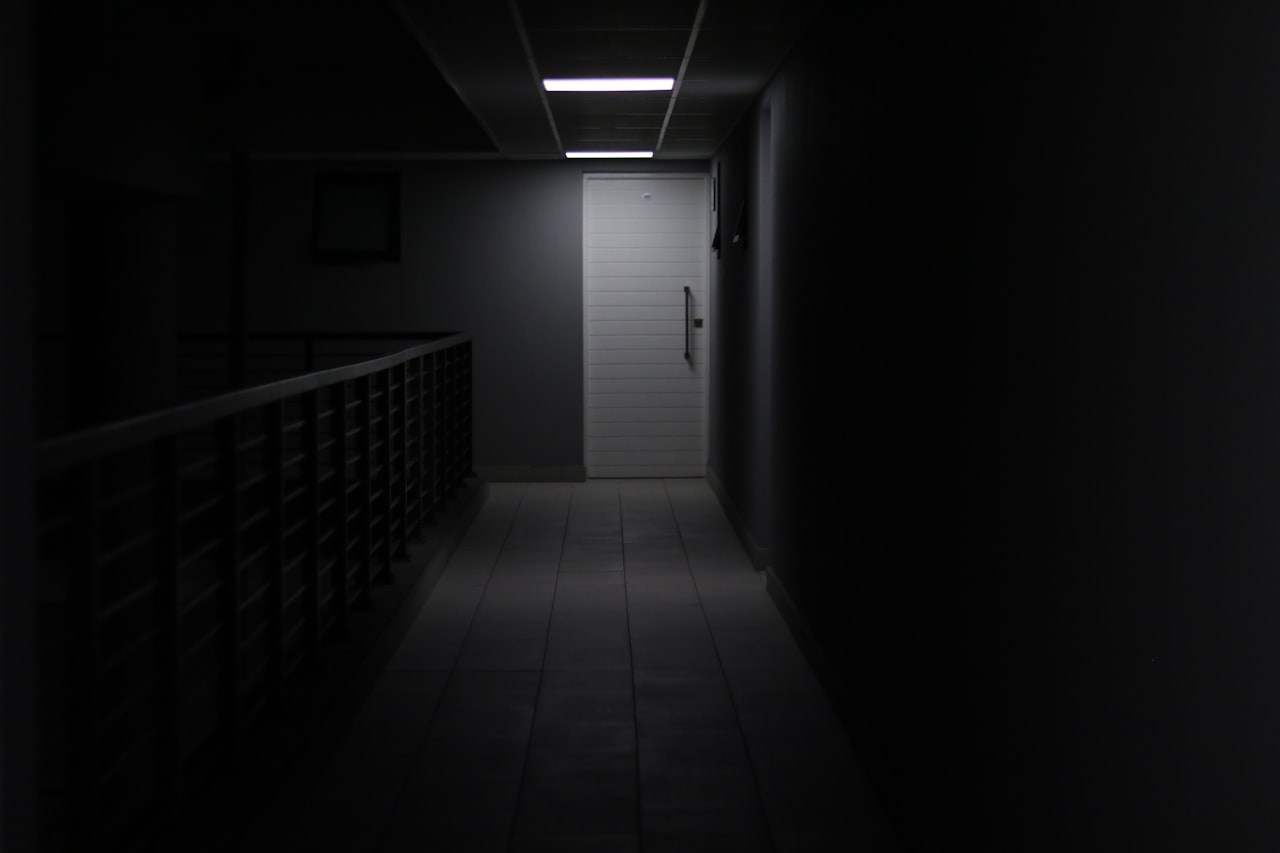 Une porte éclairée au fond d'un couloir noir — c'est dans la vision périphérique que le cerveau invente une « présence ». — Crédit : Alicia Christin Gerald / Unsplash